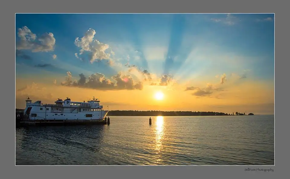 White ferry at a pier beside calm water under a low sun and radiant clouds.