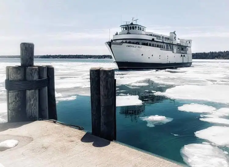 A white ferry approaches a concrete dock through broken ice, with timber pilings in the foreground.