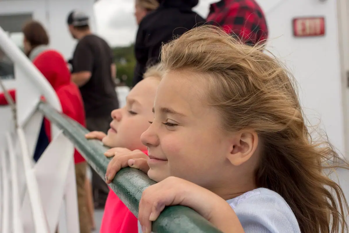 Two girls lean on a green railing while people stand behind them on a vessel deck.