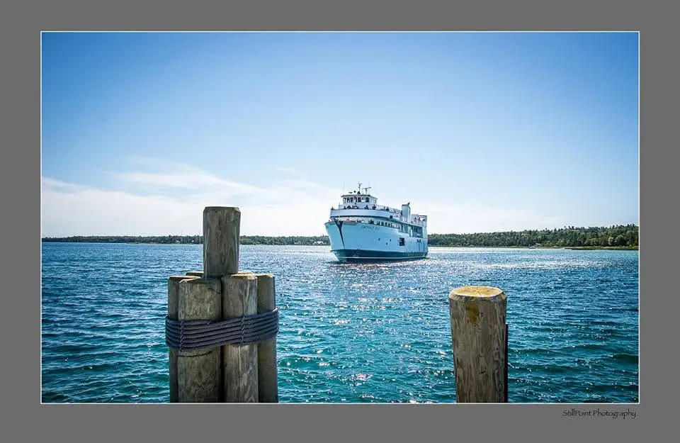 A white ferry approaches across bright blue water beyond wooden dock pilings.
