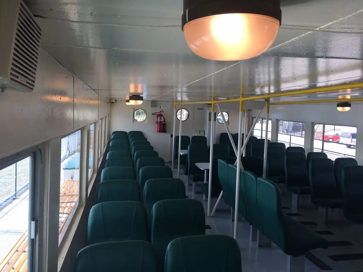 Empty passenger vessel cabin with green seats, yellow handrails, portholes, and glowing ceiling lights.