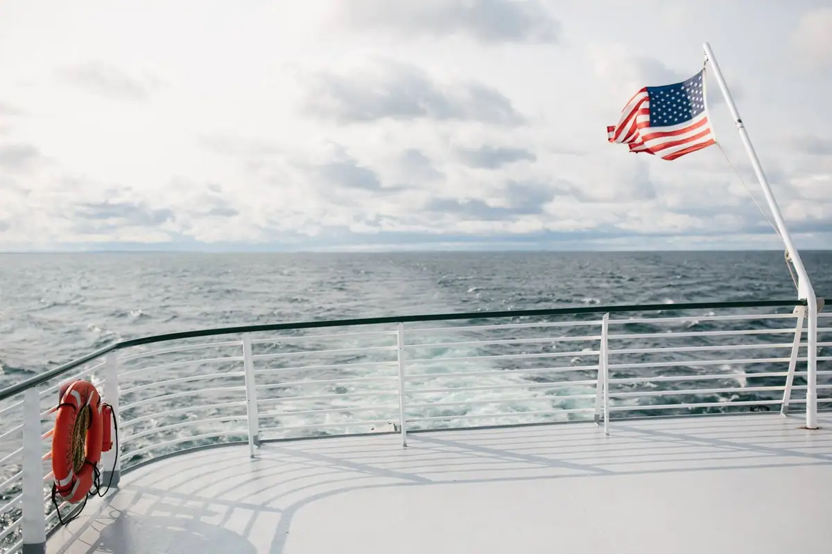 A ferry deck with white railings overlooks choppy water beneath a flying American flag.