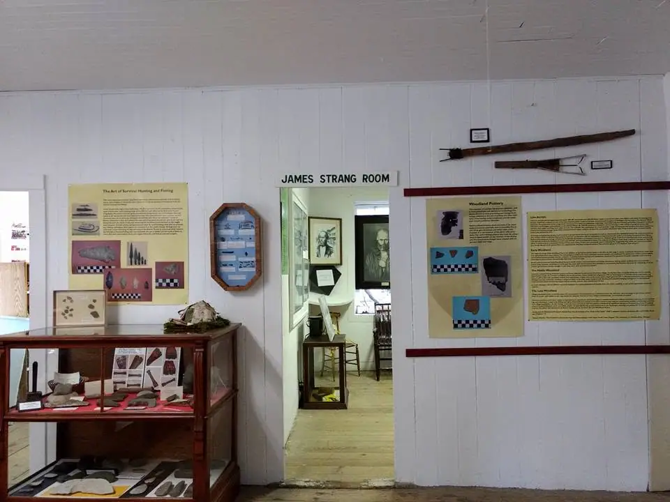 Interior exhibit room with display cases, interpretive panels, framed portraits, and mounted wooden implements.