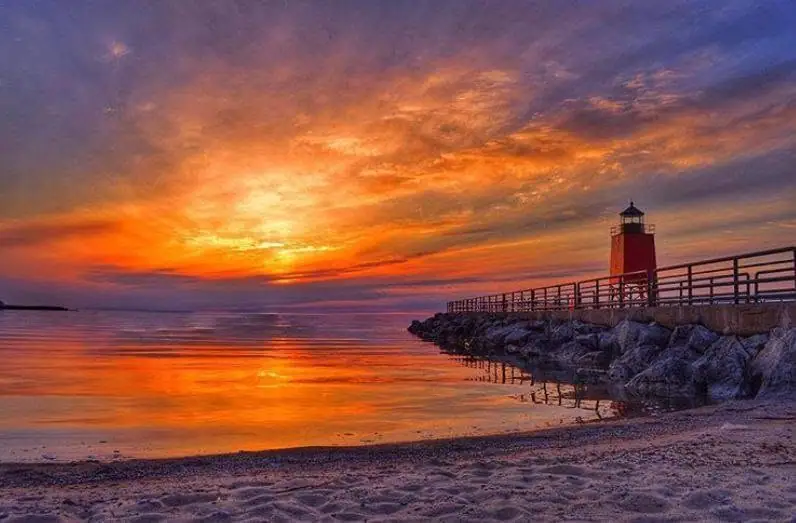 A red lighthouse stands on a rocky pier beside a beach during a vivid sunset.