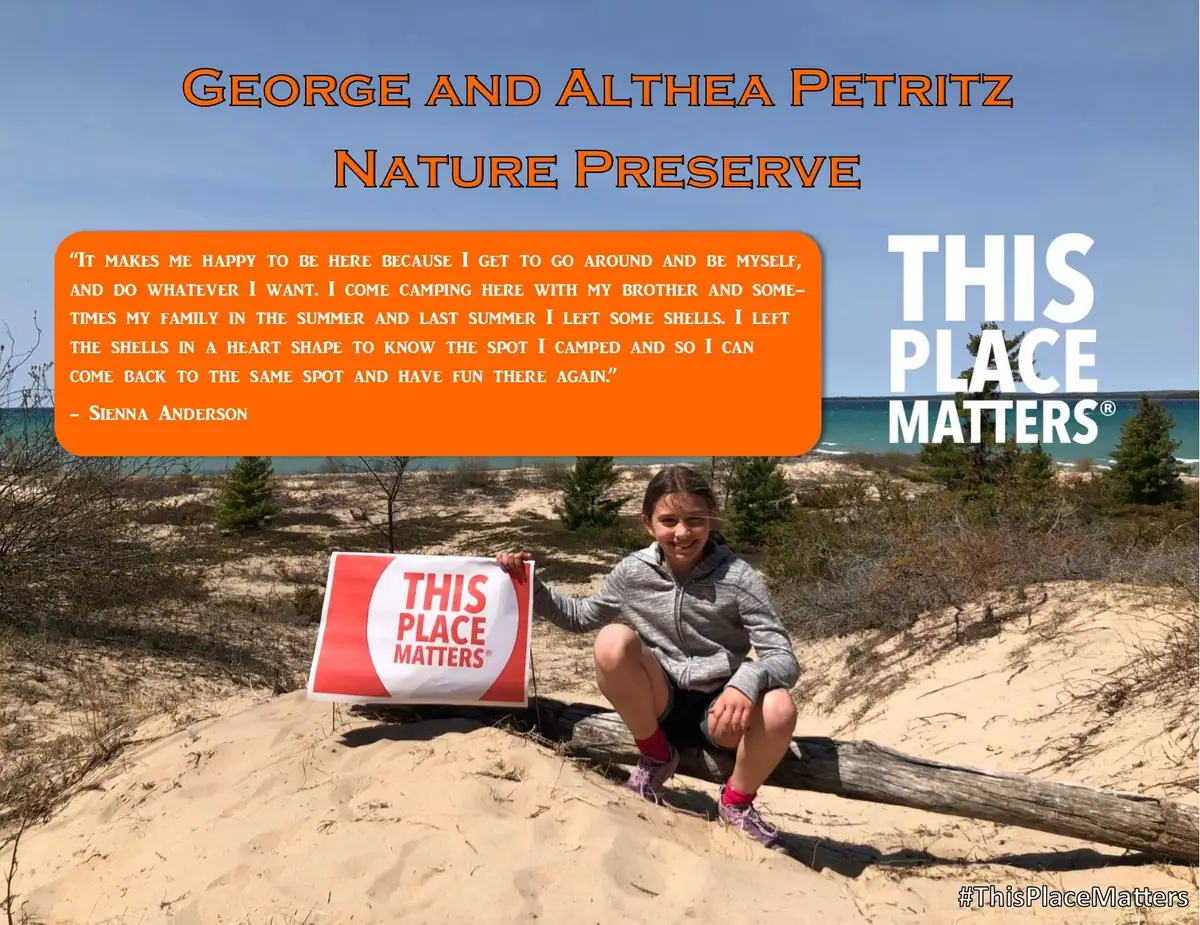 A child sits on a sandy dune beside driftwood, holding a “THIS PLACE MATTERS” sign.