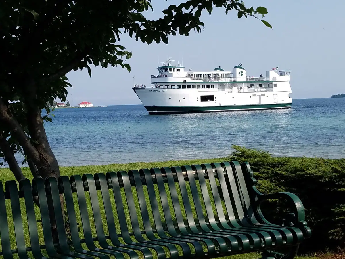 A white ferry passes offshore beyond a green metal bench beneath a leafy tree.