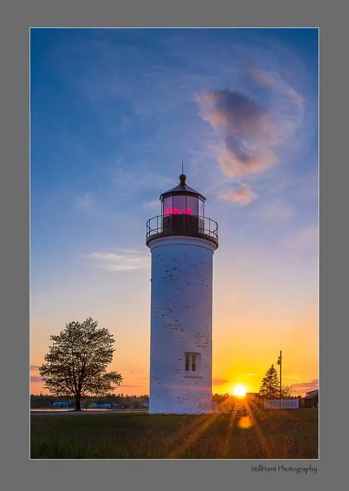 A white lighthouse stands against a colorful sunset, with a tree, fence, and small buildings nearby.