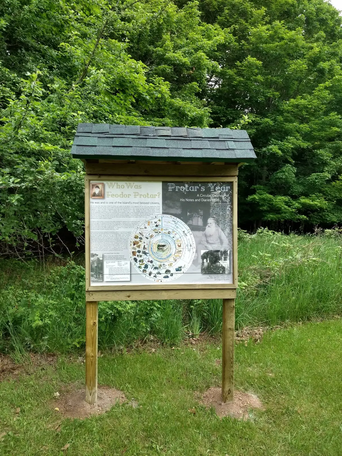A roofed wooden interpretive sign stands beside a grassy clearing at the edge of dense woods.