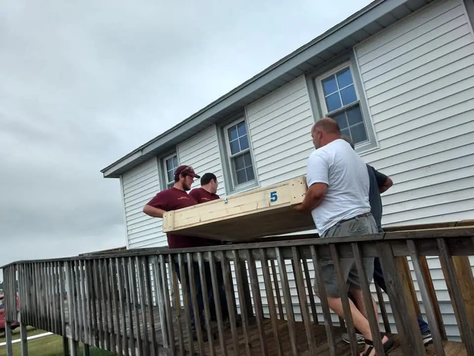 Four men carry a large wooden frame marked 5 along a raised wooden deck beside a white-sided building.