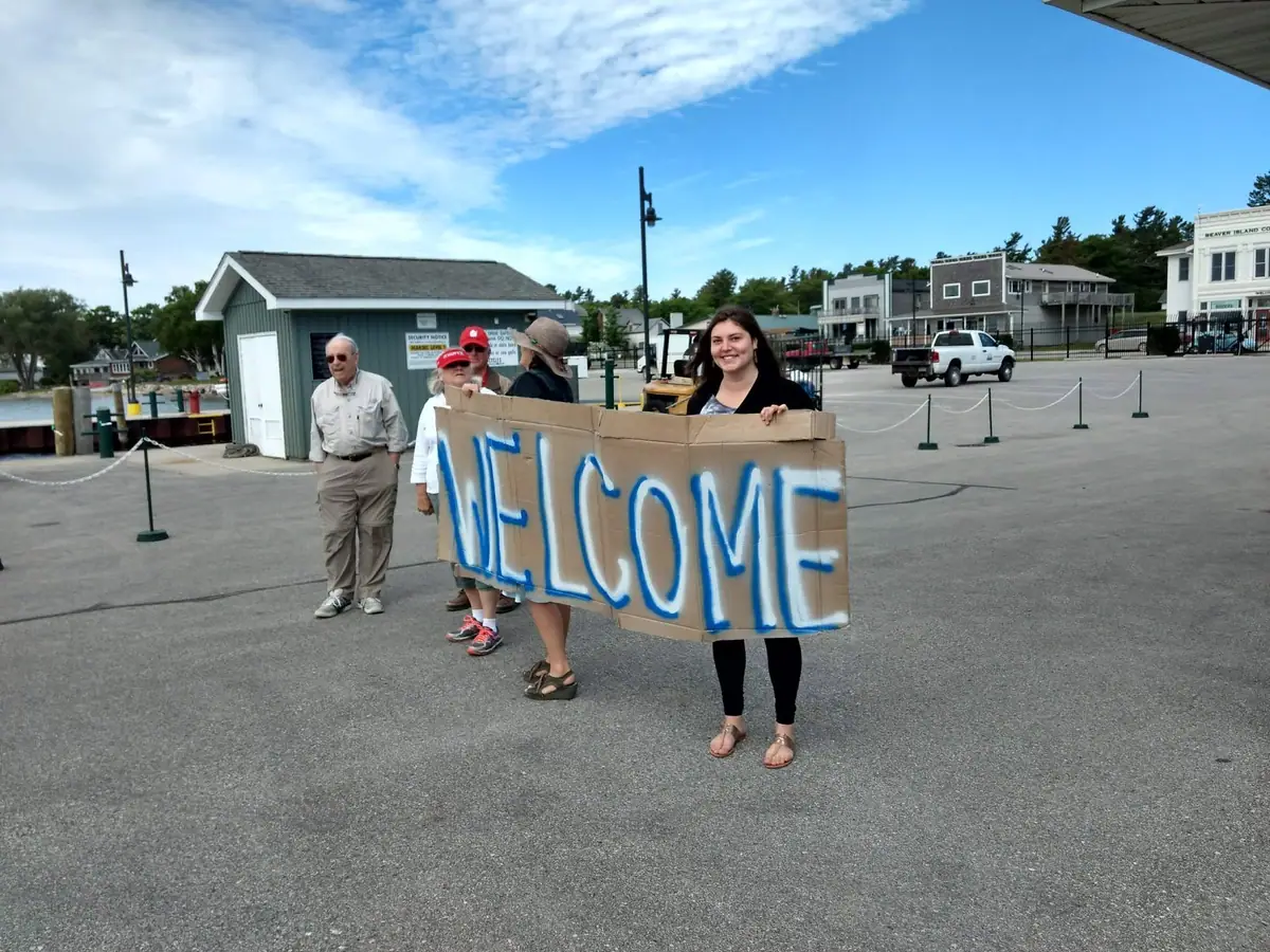 Several people hold a large hand-painted WELCOME sign in a waterfront parking area.