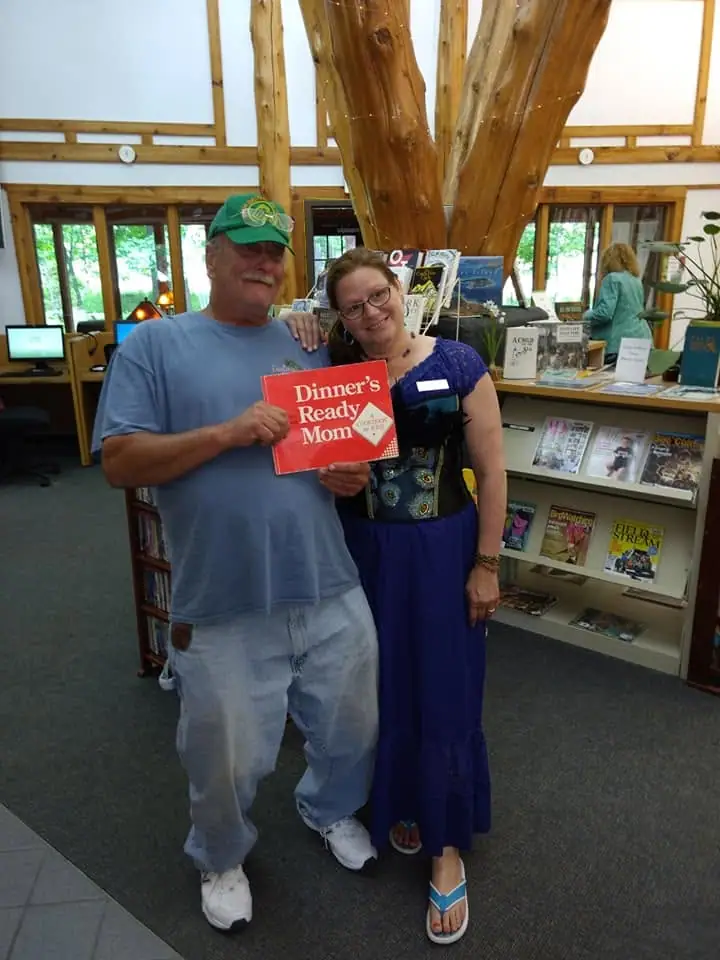 Two adults pose inside a book-filled interior while holding a red cookbook titled “Dinner’s Ready Mom.”