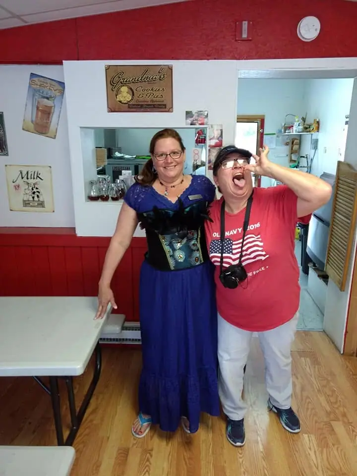 Two women pose playfully inside a brightly painted shop or service counter area.