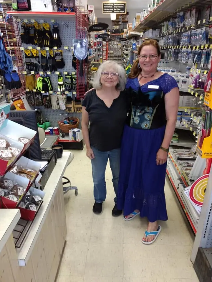 Two women pose in a hardware-store aisle surrounded by tools, gloves, shelves, and a checkout counter.