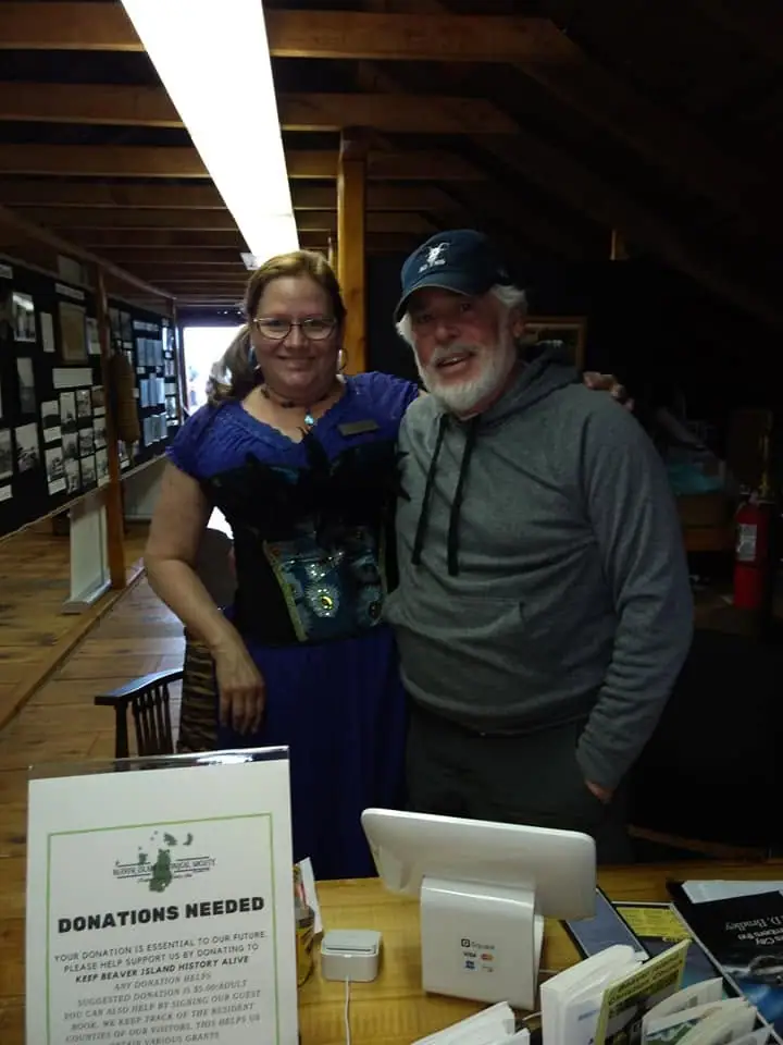 Two smiling people stand behind a donation table inside a wood-beamed museum, with exhibits and a donation sign visible.