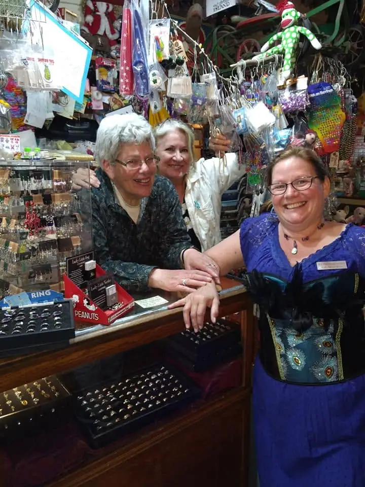 Three women pose behind a shop counter surrounded by jewelry, toys, hanging souvenirs, and colorful merchandise.