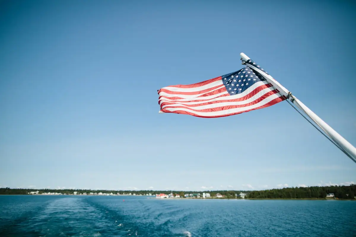 An American flag flies from an angled mast above blue water and a distant tree-lined shoreline.