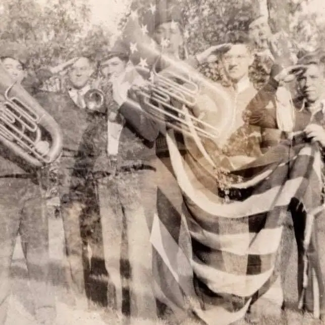 Black-and-white group portrait of men with brass instruments and a large American flag.