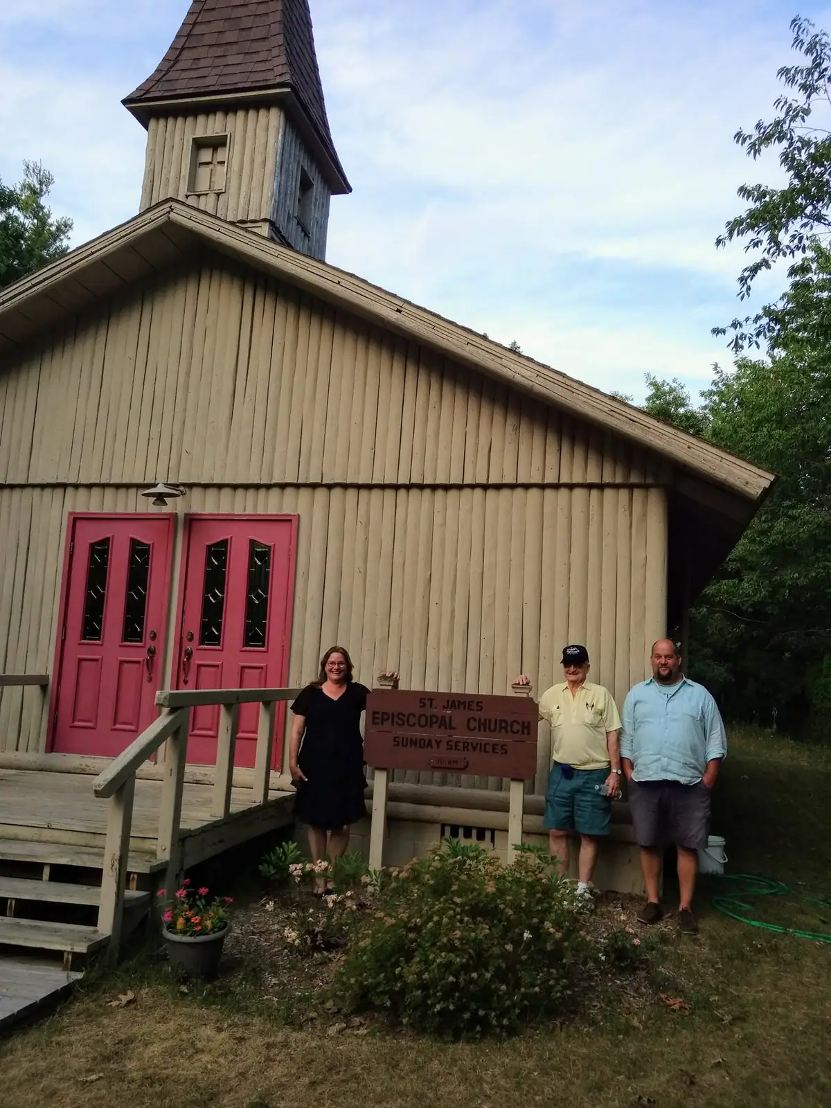 Three adults stand beside a church sign in front of a wooden church with red doors and a tower.
