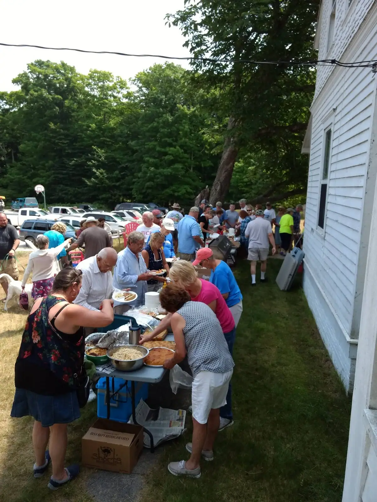 A crowd gathers around an outdoor buffet beside a white-sided building, with parked cars and trees behind them.