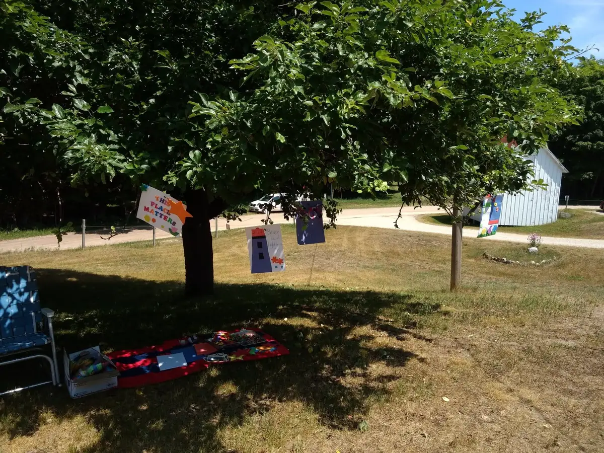 A shaded grassy picnic area with handmade signs, a blanket, a folding chair, and a white shed.