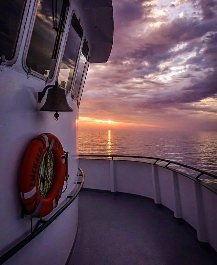 A ship’s deck with a bell and lifebuoy overlooking sunlit water beneath a colorful cloudy sky.