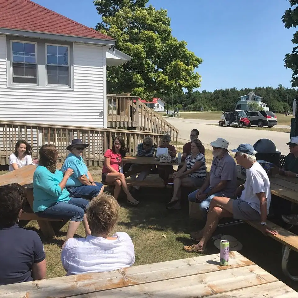 People sit at wooden picnic tables beside a white house on a sunny day.