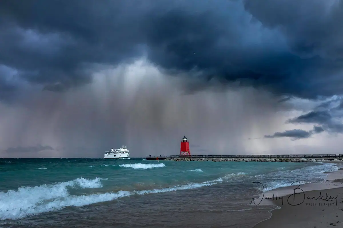 A white ferry passes a red lighthouse and pier as waves break beneath a dark, rainy storm cloud.