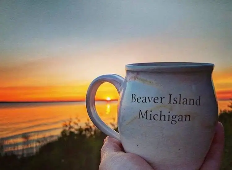 A hand holds a white mug marked “Beaver Island Michigan” against a glowing sunrise over calm water.