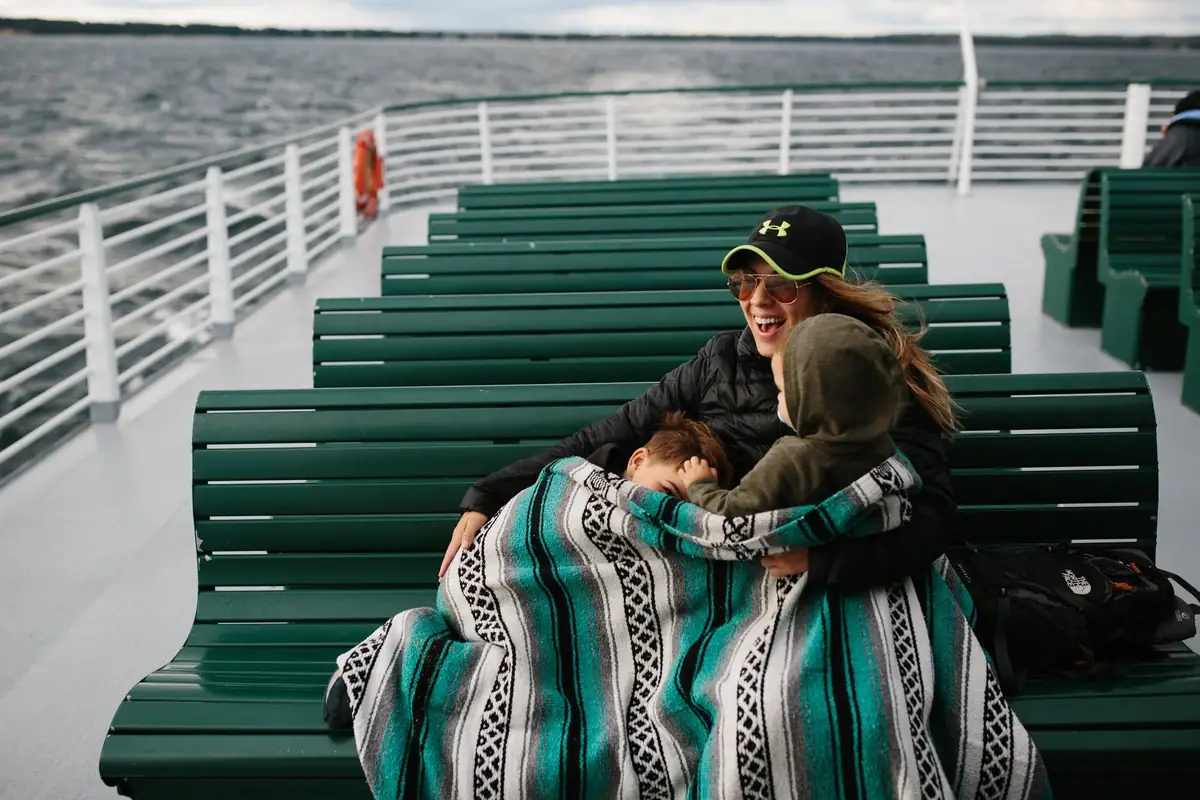 A woman embraces two children beneath a patterned blanket on a ferry deck.