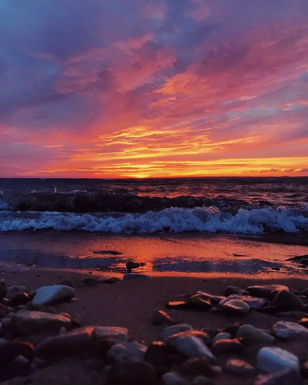 A vivid sunset glows above waves breaking on a rocky shoreline.