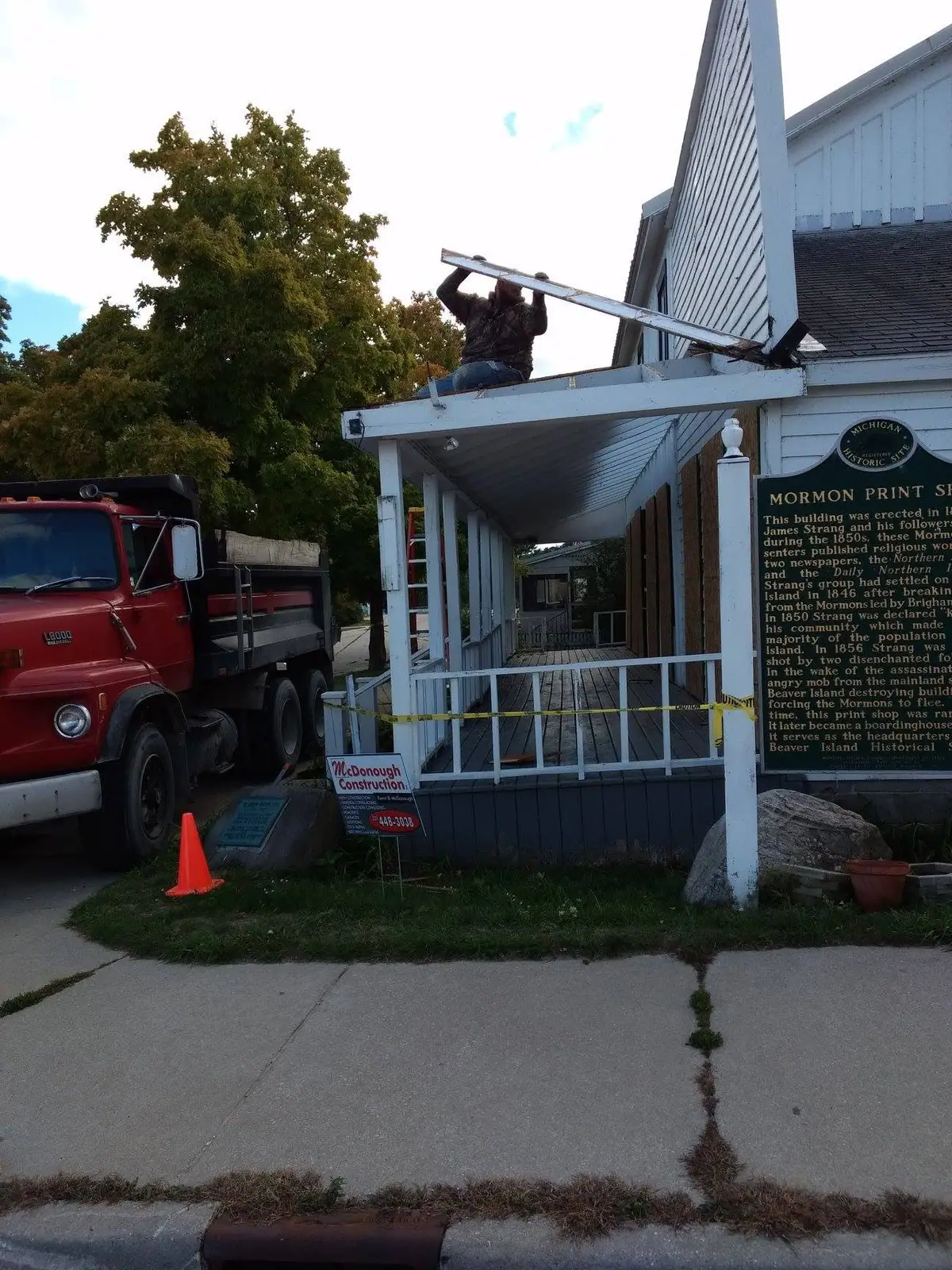 A worker installs siding on a porch beside a red dump truck and a historical marker.