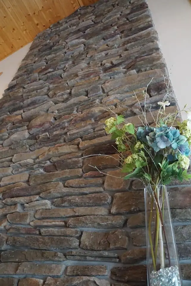 A glass vase holding blue and yellow flowers stands before a rough stone wall beneath a wooden ceiling.