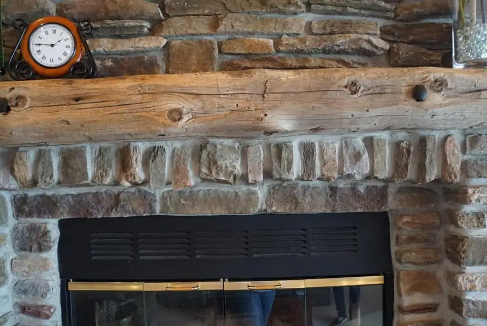 A stone fireplace has a rough wooden mantel, a round clock, and black-framed glass doors.