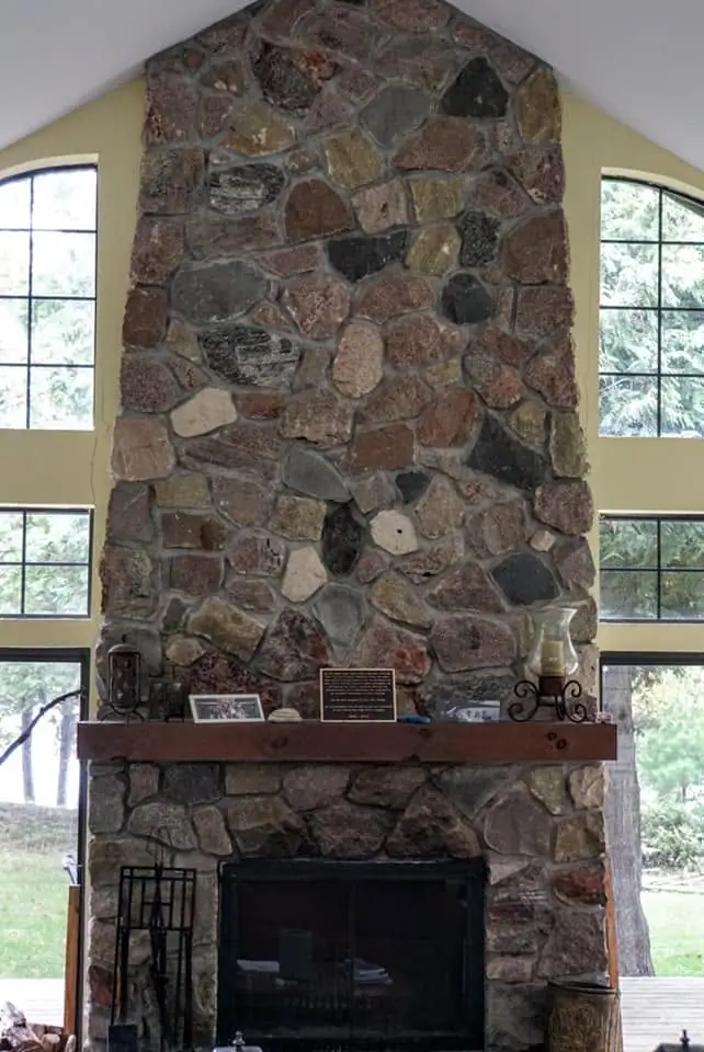 A tall stone fireplace with a wooden mantel, flanked by windows and display objects.