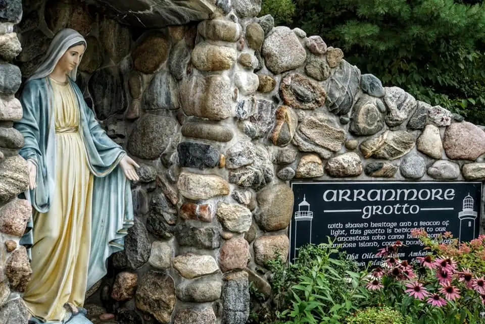 A robed statue beside a stone grotto wall, sign, and flowering plants.
