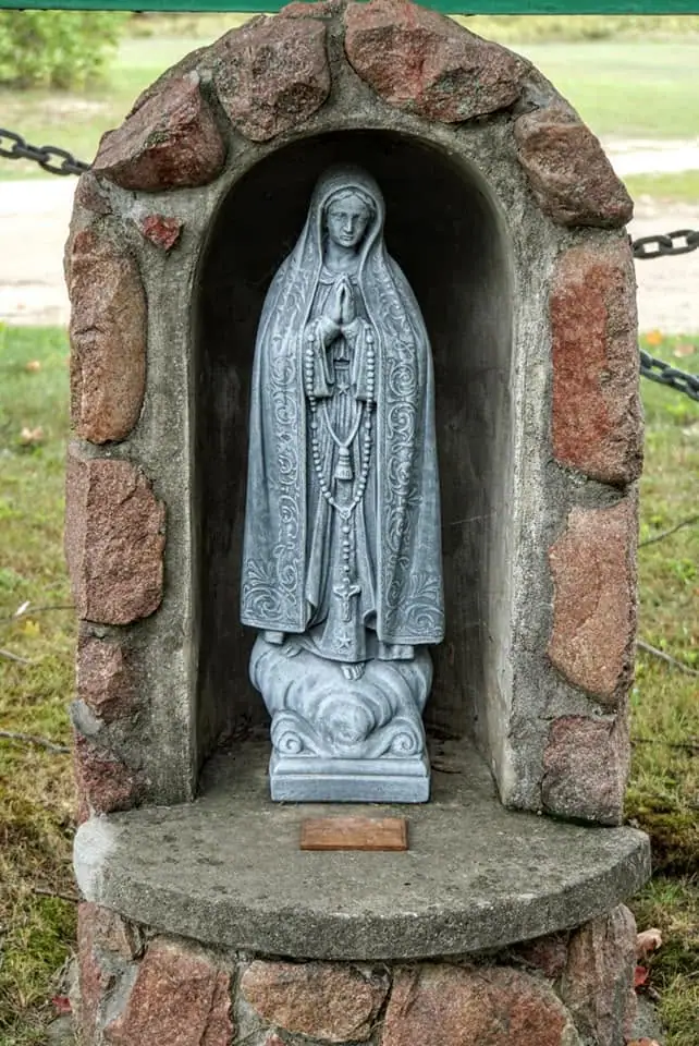 A veiled religious statue stands inside a reddish stone grotto behind a chain barrier.