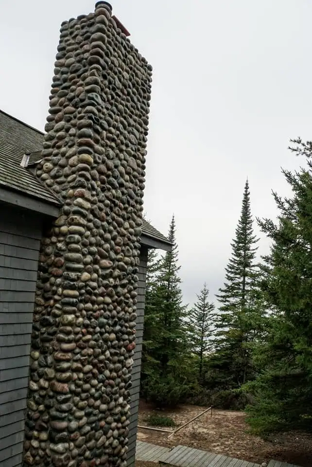 Tall rounded-stone chimney beside a shingled building and evergreen trees.
