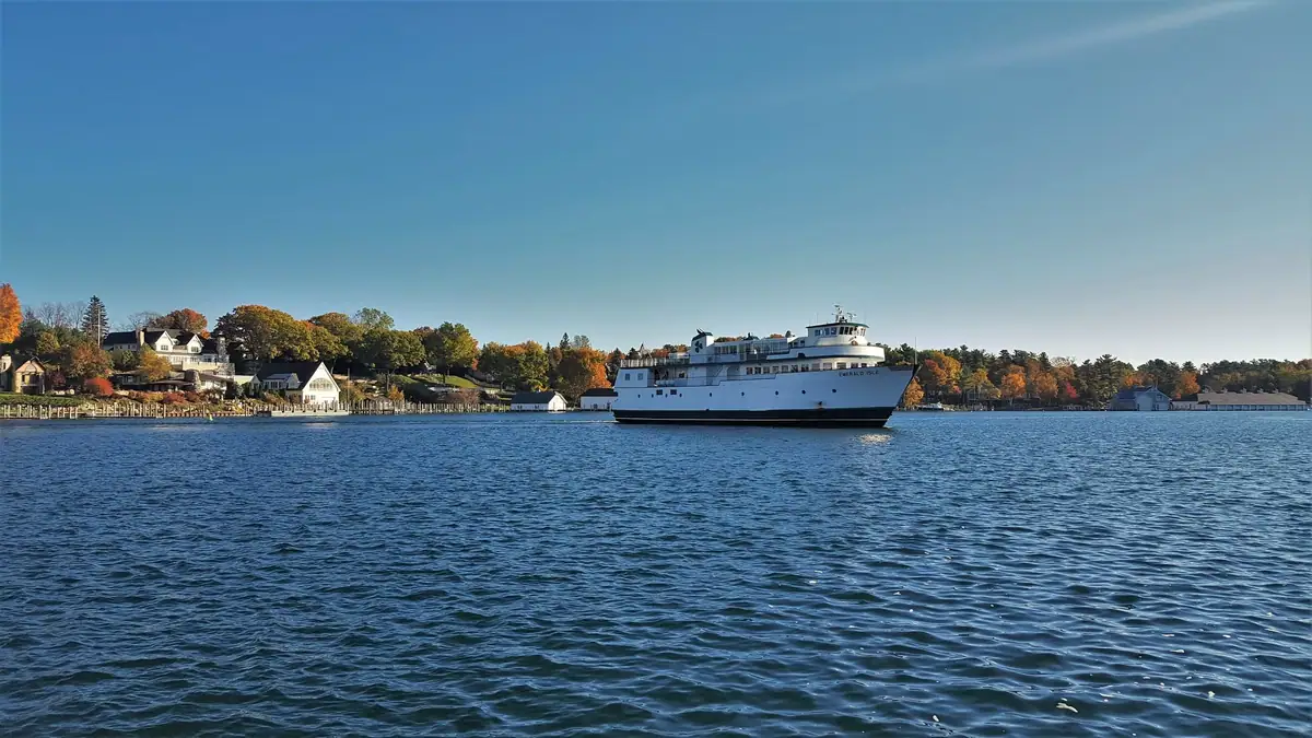 A white ferry crosses blue water beside a tree-lined shore with houses and autumn foliage.