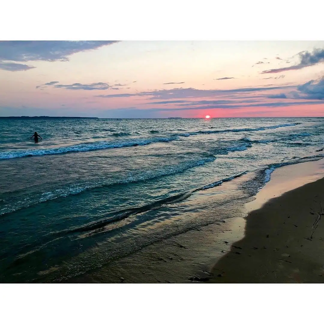 A swimmer stands in shallow waves as the sun sets over a sandy shoreline.