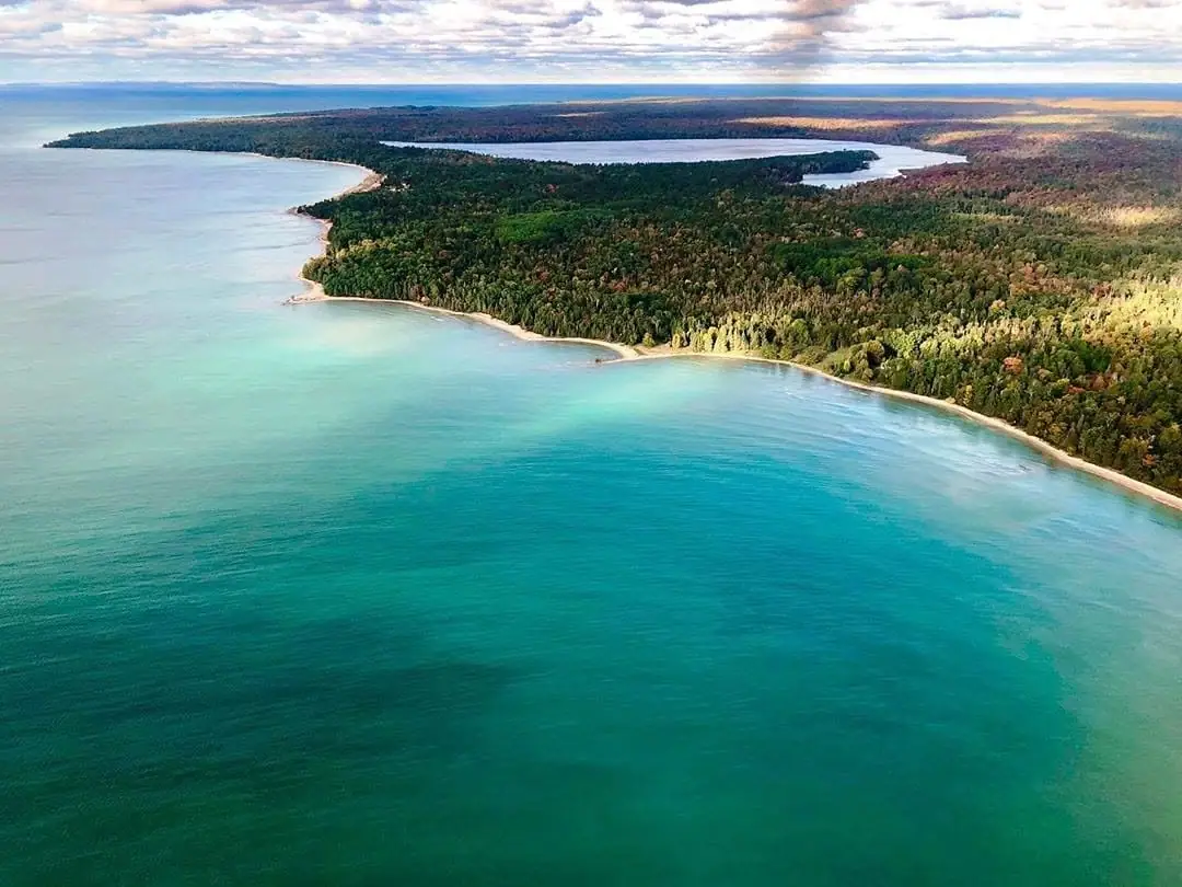 Aerial view of turquoise water curving around a forested shoreline with sandy beaches and an inland body of water.