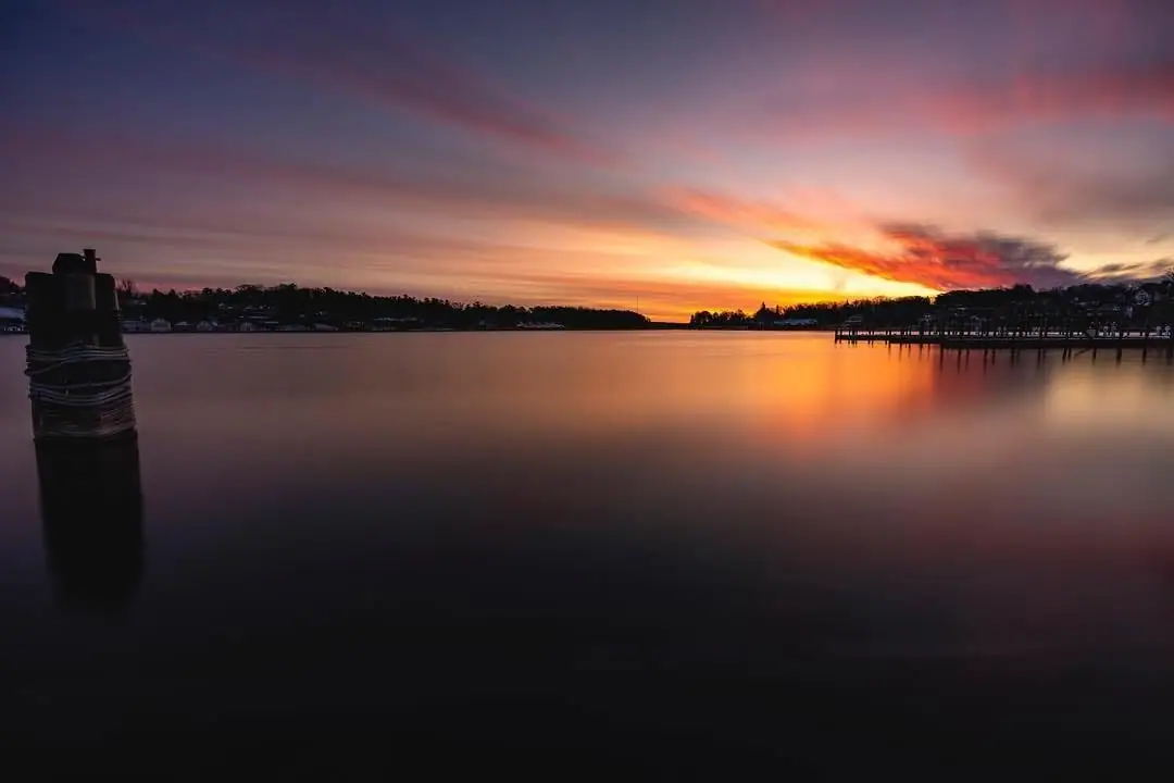 A glowing horizon reflects across calm water beside a mooring post and long wooden pier.