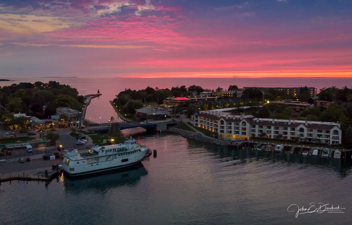 Aerial view of a harbor at sunset with a ferry, bridge, lighthouse, and waterfront buildings.