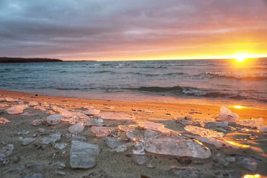 Sunset over rippling water with ice chunks scattered across a sandy shoreline.