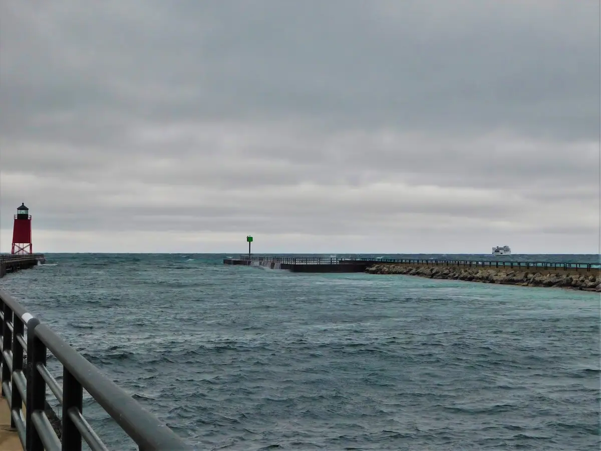 A red lighthouse and green navigation marker stand beside a breakwater in choppy water under a gray sky.