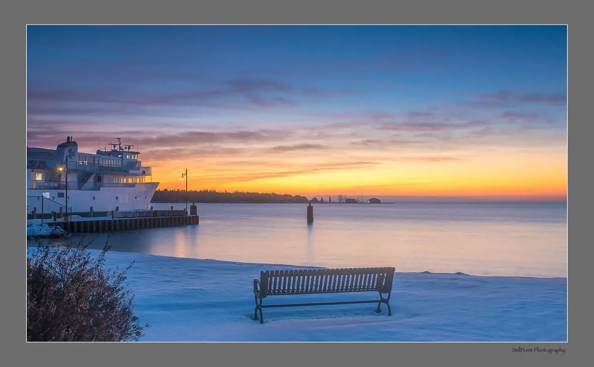 A white ferry beside a pier overlooks calm water at sunrise, with a bench on the snowy shore.