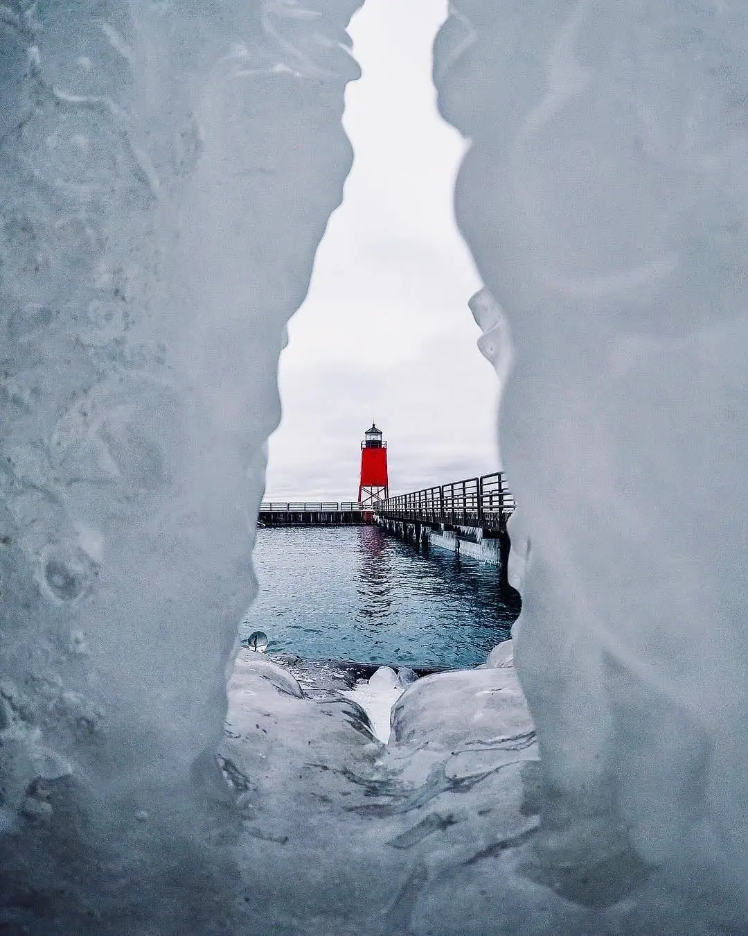 A red lighthouse and pier seen through a narrow opening in blue-white ice.