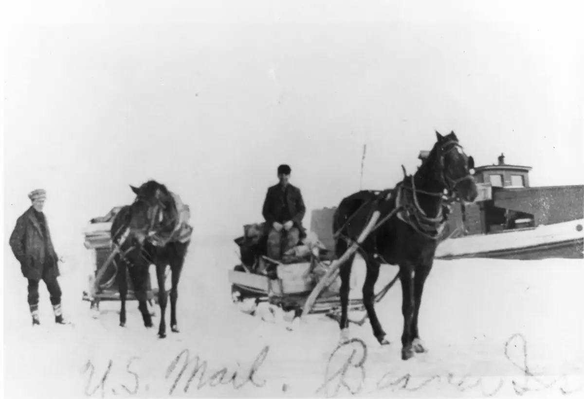 Two horses pull a mail-loaded sled through deep snow as people stand and sit nearby.