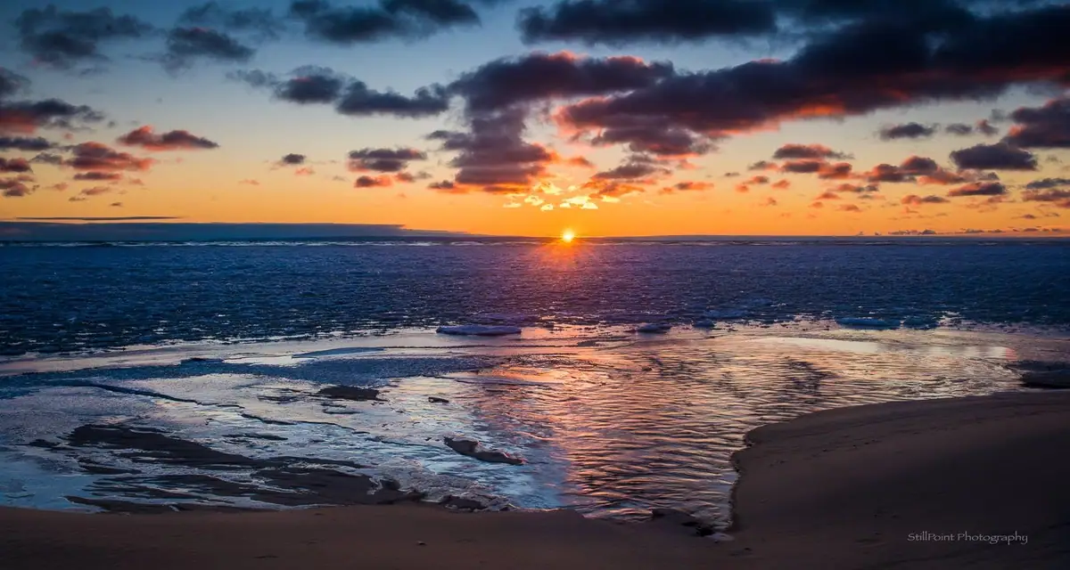 Sunrise over rippling water, with ice along a sandy shoreline beneath colorful clouds.
