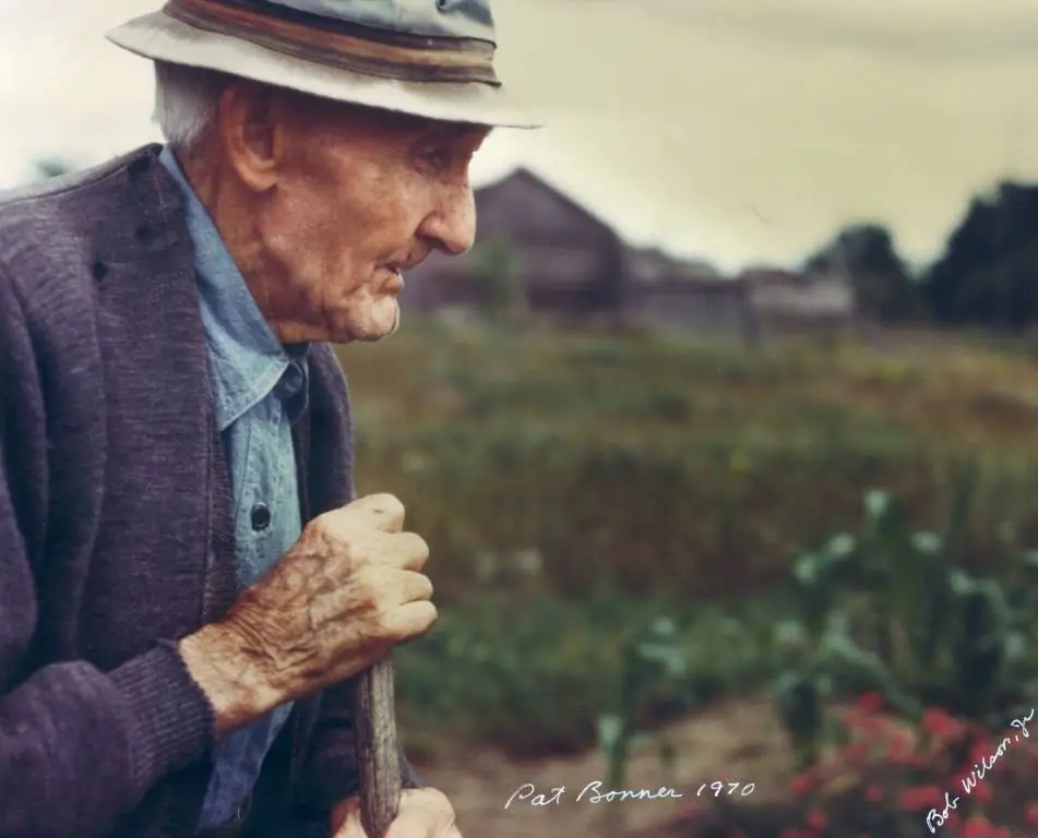 Profile portrait of an older man in a hat and cardigan, holding a walking stick beside a garden.