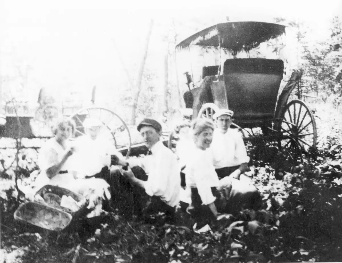 Five adults sit outdoors beside a covered horse-drawn carriage in a wooded setting.